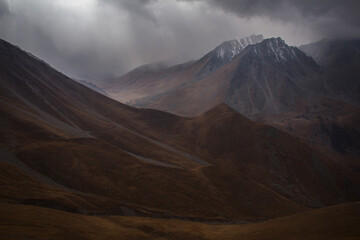 Autumn in the mountains, in a mountain valley, rain begins, the sky is in dark thick low clouds, the mountain slopes are covered with yellow grass, in the distance sunlight through the clouds