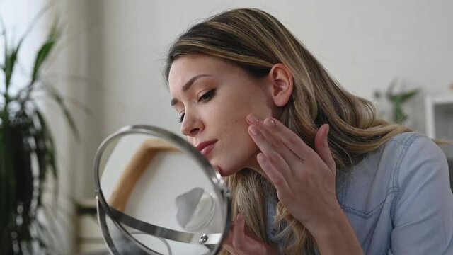 Young woman looking herself in the mirror at home. Worried about acne caused by wearing a mask. Maskne. Problems with acne.