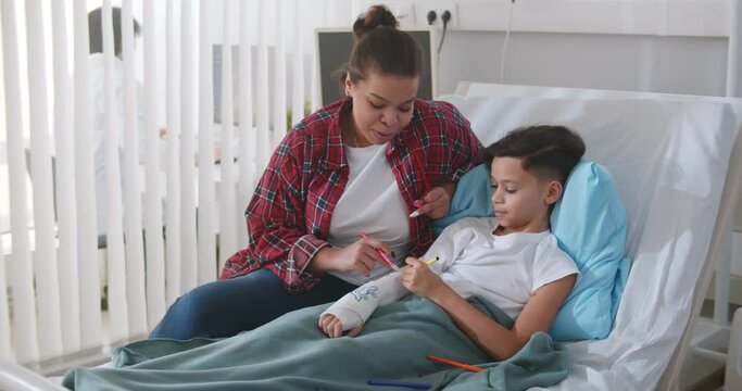 Caring african mother visiting son with broken arm in bed in hospital.