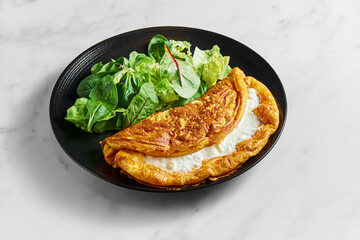 Stuffed lush creamy omelette with salad mix in black plate on white marble background