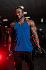 Fototapeta na wymiar Smiling black bodybuilder looks at camera with sport equipment in hands. Blue sport t-shirt and black trousers on muscular man. Closeup.