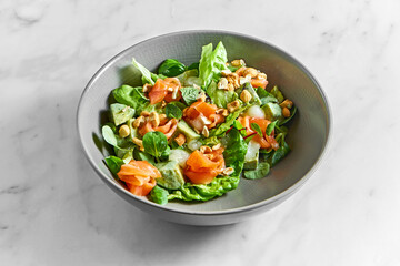 Fresh salad with salmon, delicate cheese, peanuts and mint in a gray bowl on a white marble background