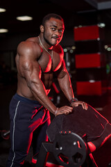 Fototapeta na wymiar Pumped up shirtless african american sportsman in modern gym with up to date sport equipment. Cute black man under dramatic red law key lightning. Closeup.