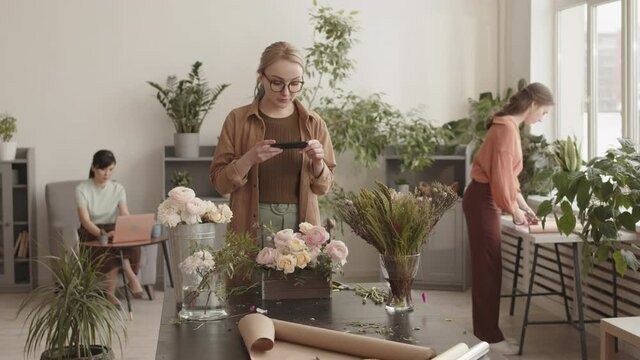 Steadicam of blonde Caucasian woman standing by table in plant shop, taking pictures of flowers arrangement in wooden box, female colleagues working on background