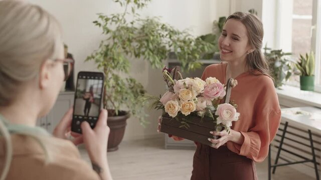 Medium over shoulder of blond-haired Caucasian woman taking picture of young female friend, holding wooden basket with flowers arrangement, standing in plant shop, then showing her picture