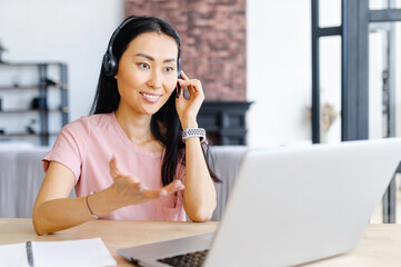 Asian businesswoman having call via laptop application, sitting at the desk, showing with the hand, holding microphone with her hands, checking documents, new normal working from home, online training