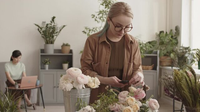 Medium shot of young blond-haired Caucasian woman standing in plant shop, taking pictures of flowers arrangement in wooden box, female colleague working with laptop on background
