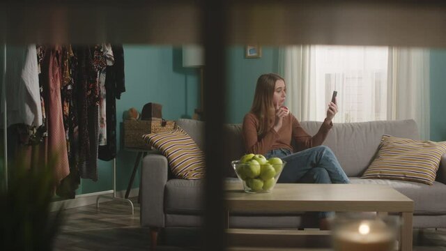 Young woman in blue jeans and brown sweater sits on beige sofa on window background, bites apple and talks on video call. On coffee table is glass jar with green apples. Camera dollies horizontally