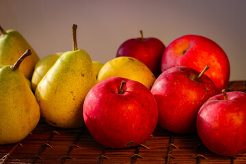 Several ripe yellow pears and red apples stand on a wicker basket illuminated by rays of the sun