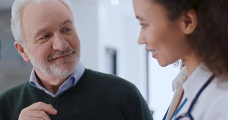 Portrait multiethnic female doctor in white medical coat and happy senior male patient talk about great news from his health report standing in hospital. Mixed race nurse and elderly caucasian man - Powered by Adobe