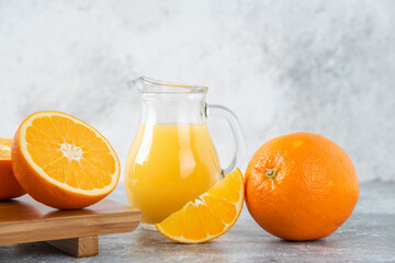 A glass pitcher of juice with fresh orange fruits on stone background