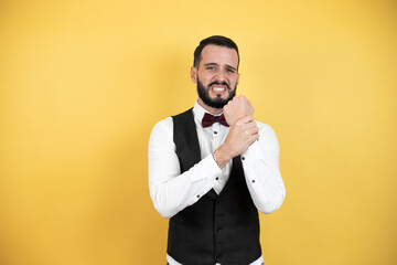 Young man with beard wearing bow tie and vest suffering pain on hands and fingers, arthritis inflammation