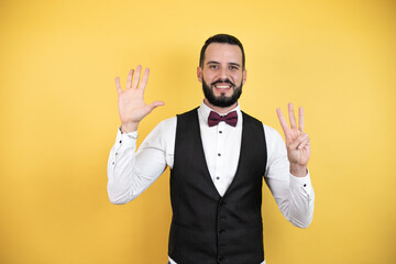 Young man with beard wearing bow tie and vest showing and pointing up with fingers number eight while smiling confident and happy