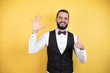 Young man with beard wearing bow tie and vest showing and pointing up with fingers number six while smiling confident and happy