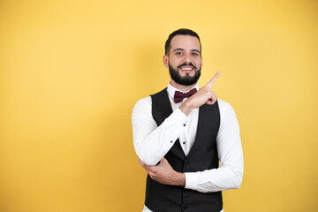 Young man with beard wearing bow tie and vest amazed and pointing with hand and finger to the side