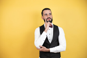 Young man with beard wearing bow tie and vest serious face thinking about question with hand on chin, thoughtful about confusing idea
