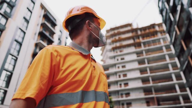 Topic is health protection in production and construction. Caucasian male worker in orange uniform and protective respirator kn 95 puts on construction orange hard hat and poses at construction site