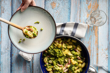 Top view on fresh prepared broccoli with gnocchi in casserole dish on the wooden table - hand of unknown person putting homemade healthy food in a plate over the table in bright light with copy space