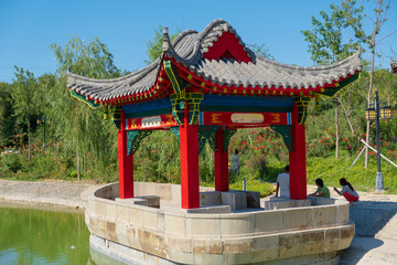 Chinese style pavilion with upturned eaves on roof in the park near the rose garden with family peoples walking and resting near the water pond