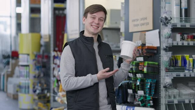 Portrait of confident smiling man advertising paint in hardware store. Brunette Caucasian seller with brown eyes pointing at can looking at camera. Choice of repair supplies.