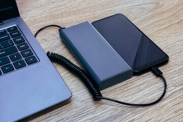 Smartphone is being charged from a power bank. Portable charger with laptop on a wooden table.