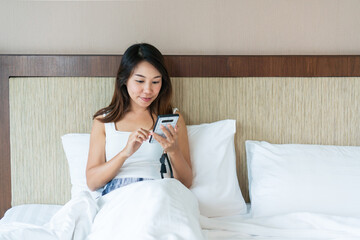 Young happy Asian woman sitting on bed and using mobile phone. Young brunette girl relaxing at home in a leisure time.