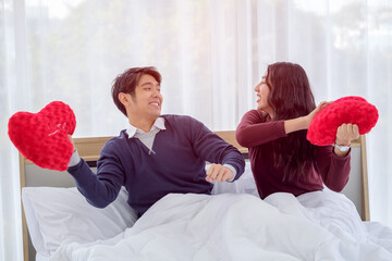 Smiling young asian couple on white bed with red heart-shaped pillow,  Valentine's day and happiness concept.