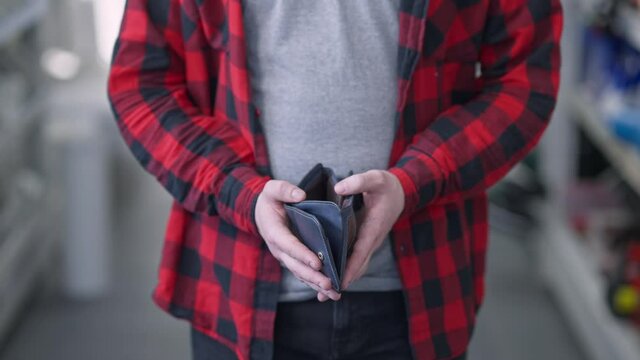 Close-up of male hands opening empty wallet. Unrecognizable Caucasian man standing between blurred rows with goods in shop having no money. Consumerism and poverty.
