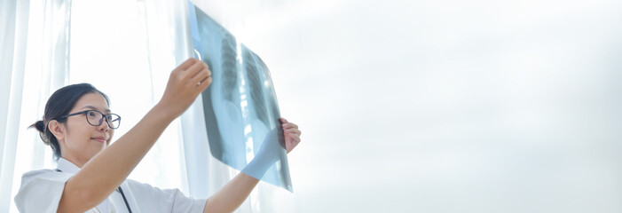 Smiling Asian Young Female doctor holding looking and examining X-ray film of her patient in medical room of hospital. Banner and copy space