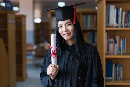 A young happy Asian woman university graduates in graduation gown and cap wears a face mask holds a degree certificate to celebrate her education achievement on the commencement day. Stock photo