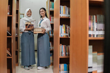 two female students in high school uniforms carry several books standing between the bookshelves