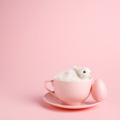 Easter bunny rabbit in a teacup with painted egg on pastel pink background. Creative Easter holiday concept with copy space. Minimal spring holiday greeting card.
