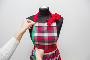 Young adult woman hands measuring kitchen apron with green measuring tape on mannequin isolated on light gray wall background. Right size. Closeup. Handmade work. Feminine hobby. Point of view shot.
