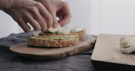man making ciabatta bruschettas with mozzarella and pesto on olive board with window on backgorund