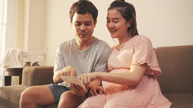 Asian couple husband and pregnant woman watching ultrasound film happy feeling on sofa at home, New family baby born concept.