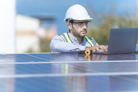 Engineer Man Inspects Construction Of Solar Cell Panel Or Photovoltaic Cell By Laptop Device. Industrial Renewable Energy Of Green Power. Factory Worker Working On Tower Roof And Skyscraper City.