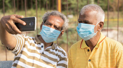 elderly friends with medical face mask taking selfie while sitting at park - concept of happy senior people friendship and meetup after lockdown due to coronavirus covid-19 pandemic. - Powered by Adobe