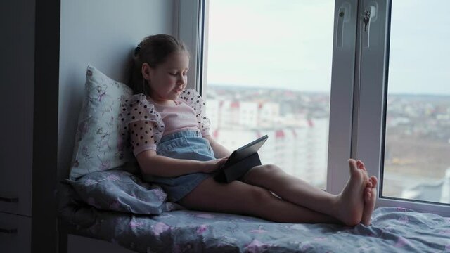 Children and technology. An attractive girl sits near the window and looks at the tablet. She smiles and laughs. The city is visible outside the window