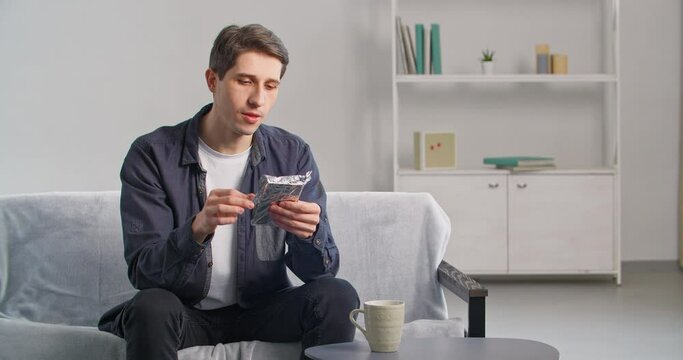 Young man sits on sofa at home holding wrapped chocolate bar in his hands opens dessert sniffs aromatic smell of cocoa bites expresses pleasure from taste enjoys sweetness during breakfast or lunch 
