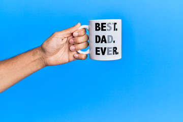 Hand of hispanic man holding best dad ever coffee cup over isolated blue background.