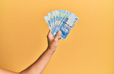 Hand of hispanic man holding south africa rand banknotes over isolated yellow background.
