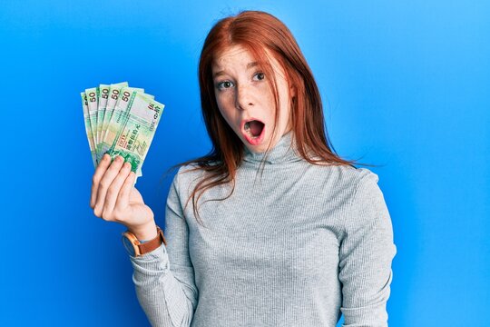 Young red head girl holding 50 hong kong dollars banknotes scared and amazed with open mouth for surprise, disbelief face