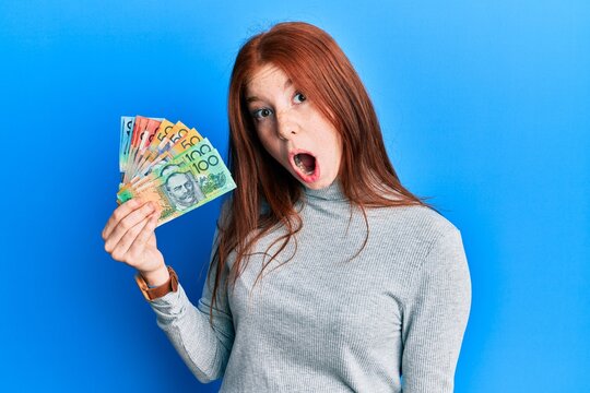 Young red head girl holding australian dollars scared and amazed with open mouth for surprise, disbelief face