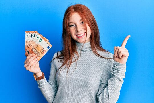 Young red head girl holding bunch of 50 euro banknotes smiling happy pointing with hand and finger to the side