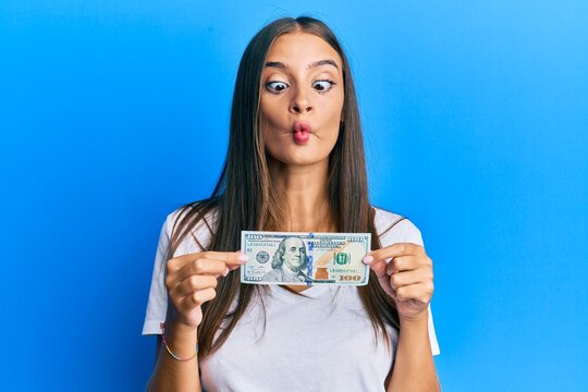 Young hispanic woman holding 100 dollar banknote making fish face with mouth and squinting eyes, crazy and comical.