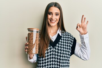 Beautiful brunette young woman holding jar of healthy whole grain cereals doing ok sign with fingers, smiling friendly gesturing excellent symbol