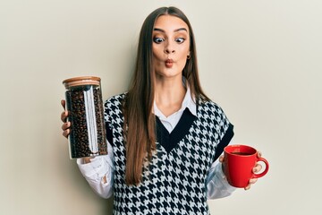 Beautiful brunette young woman holding jar with coffee beans and drinking coffee cup making fish...