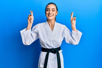 Beautiful brunette young woman wearing karate fighter uniform with black belt gesturing finger crossed smiling with hope and eyes closed. luck and superstitious concept.