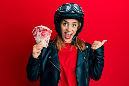Hispanic young woman wearing motorcycle helmet holding 20 shekels banknotes pointing thumb up to the side smiling happy with open mouth