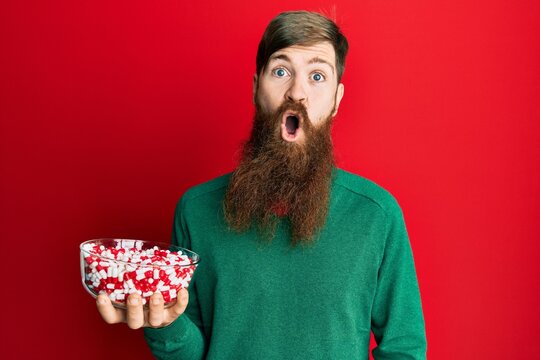 Redhead man with long beard holding bowl full of pills scared and amazed with open mouth for surprise, disbelief face